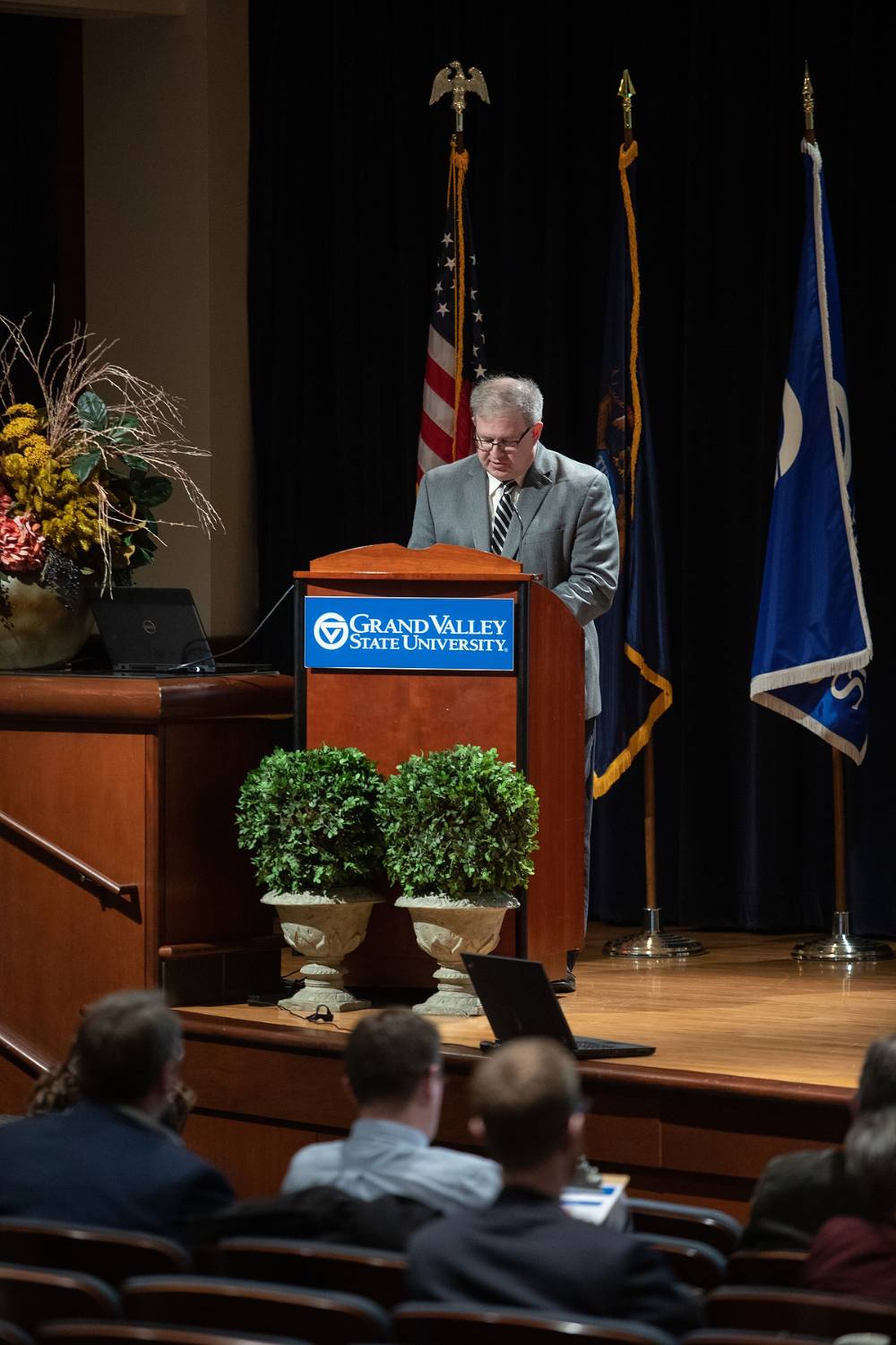 Man glancing down at a GVSU podium on stage, with audience members sitting below the stage